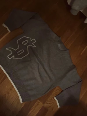 Grå stickad tröja med text - Säljer en grå stickad tröja med vit text på bröstet och coolt tryck på ryggen där det står 'buy happiness'. Tröjan har långa ärmar, ribbade muddar och kontrasterande vita detaljer vid ärmslut och nederkant. Perfekt för dig som gillar streetwear vibes.