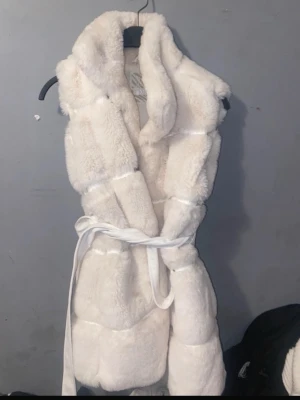 Vit pälsväst från MISU med bälte - Säljer en fluffig och lyxig vit pälsväst från MISU. Västen har en bred krage och knyts i midjan med ett matchande bälte för extra snygg passform. Perfekt att slänga över en hoodie eller stickad tröja när det är kallt ute.