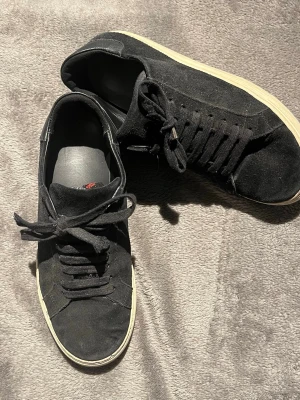 Axel Arigato skor Svart i storlek 40 - Säljer ett par svarta sneakers från Axel Arigato med ovandel i mocka och detaljer i skinn vid hälen. Skorna har snörning och en vit, platt sula som ger en clean look. Perfekta för dig som gillar stilrena och klassiska sneakers. Hör gärna av dig ifall att du har frågor.