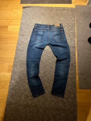 Nudie Jeans blå jeans, storlek 31/30 - Säljer ett par klassiska blå jeans från Nudie Jeans i storlek 31/30. Jeansen har en snygg tvätt med lätt slitning, raka ben och fem fickor. Gula kontrastsömmar och logga på bakfickan.