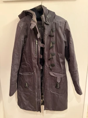 Mörkgrå parkajacka med huva från Elvine - Säljer en mörkgrå parkajacka från Elvine med mysigt svart foder. Jackan har huva, stora fickor framtill, snygga toggle-knappar och dragkedja. Perfekt för kalla dagar och har en längre modell som täcker bra. Stilsäker och funktionell jacka för vintern.