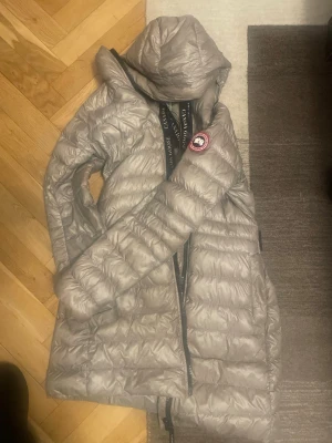Ljusgrå dunjacka från Canada Goose - Säljer en lång, ljusgrå dunjacka från Canada Goose med huva och klassisk logga på ärmen. Jackan har quiltad design, dragkedja framtill och tryckknappar vid fickorna. Perfekt för kalla dagar och riktigt snygg till vinterlooken.