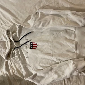 Vit hoodie från Gant med logga - Säljer en vit hoodie från Gant med broderad logga och amerikanskt sköldmärke på bröstet. Tröjan har huva med svarta snören och en stor magficka framtill. Perfekt för dig som gillar klassisk och sportig stil.