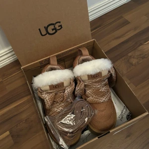 UGG Lowmel Sneakers ”Chestnut” - UGG Lowmel sneakers i brun mocka och textil med grov sula och breda, mönstrade snören. Skorna har vit fluffig fårullskant och UGG-logga på sidan och plösen. Perfekta för kalla dagar med sin varma insida och snygga outdoor-vibe.