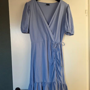 Ljusblå omlottklänning Gina Tricot - Supersöt ljusblå omlottklänning från Gina Tricot med puffärmar, v-ringning och knyt i midjan. Klänningen har volang nertill och lätt rynkdetalj på sidan. Perfekt för sommarens alla tillfällen!