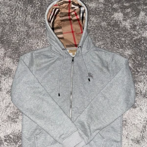 Grå Burberry hoodie med dragkedja - Snygg grå hoodie från Burberry med klassiskt rutigt foder i huvan. Tröjan har dragkedja framtill, snörning i huvan och broderad logga på bröstet. Tillverkad i mjuk bomullsmix för extra komfort. Perfekt för dig som vill ha en stilren och lyxig look.