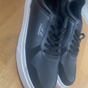 Svarta sneakers från Vitale  - Snygga svarta sneakers från Vitale med vit sula och klassisk design. Skorna har snörning och är gjorda i syntetmaterial med stilren logga på sidan. Perfekta för dig som gillar en clean och modern look.