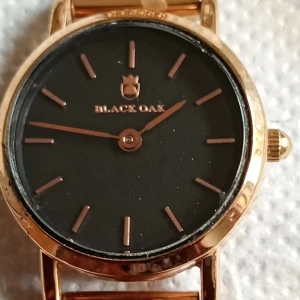 Black Oak Gerardii Petite klocka guld - Stilren klocka från Black Oak, modell Gerardii Petite. Klockan har svart urtavla med guldiga markeringar och boett i guldton. Armbandet är ett tunt meshband i guld, vilket ger en modern och elegant look. Vattentålig upp till 3 ATM. Fullt fungerande 