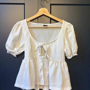 Vit blus med puffärm från Gina Tricot - Supersöt vit blus från Gina Tricot i storlek XS. Blusen har puffärmar, volang nertill och knytning framtill.