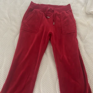 Röda mjukisbyxor från Juicy Couture - Röda mjukisbyxor från Juicy Couture i mjuk velour med bred resår och snörning i midjan. Stora fickor fram och bak med tryckknappar och broderad JC-logga på ena fickan. Perfekta för en chill och trendig look.