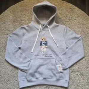 Grå hoodie Polo Bear Ralph Lauren - Snygg grå hoodie från Polo Ralph Lauren med ikoniska Polo Bear-trycket framtill. Tröjan har huva med vita snören och en stor magficka. Mjuk bomullsblandning och klassisk passform, perfekt för chill dagar. Accepterar bud.