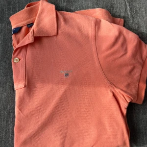 Korallfärgad pikétröja från GANT - Snygg korallfärgad pikétröja från GANT med klassisk krage och två knappar framtill. Tröjan har broderad logga på bröstet och är tillverkad i mjuk bomull som andas. Perfekt för dig som gillar stilrena och sportiga plagg.