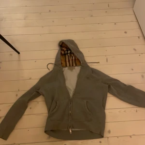 Grå hoodie från Burberry med dragkedja - Säljer en grå hoodie från Burberry med klassiskt rutigt foder i huvan. Tröjan har dragkedja framtill, två fickor och snörning i huvan. Perfekt för dig som vill ha en stilren och lyxig look med en touch av ikoniskt mönster.