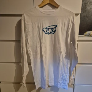 Vit långärmad tröja från Vans - Vit långärmad tröja från Vans med blå logga framtill i streetstil. Tröjan har rund halsringning och är gjord i mjuk bomull, perfekt för en avslappnad look. Passar dig som gillar enkel och clean stil med en touch av skate.
