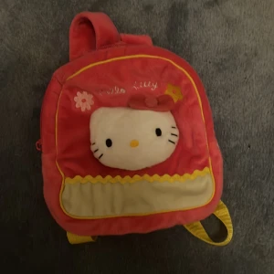 Hello Kitty rosa ryggsäck i plysch - Supergullig rosa ryggsäck från Hello Kitty i mjuk plysch med broderade blommor och ett stort Hello Kitty-ansikte framtill. Väskan har gula axelband och dragkedja, samt gult innerfoder. Perfekt för dig som älskar söta accessoarer och vill sticka ut. Den är väldigt liten däremot!