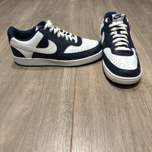 Nike Court Vision Low sneakers blå/vit - Snygga Nike Court Vision Low sneakers i marinblått och vitt. Klassisk låg modell med vit sula, perforerad tå och tydlig Nike Swoosh på sidorna. Ovandel i syntet och skinn, med vita snören och bekväm stoppning runt ankeln. Perfekta för en clean och sportig look. Använda 5-6 gånger