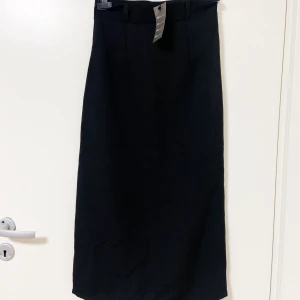 Svart långkjol från Awesome Products - Svart långkjol från Awesome Products med stilren och rak siluett. Kjolen har bälteshällor i midjan och en diskret slits baktill för extra rörelsefrihet. Materialet är mjukt och har en lätt glansig finish som ger en snygg look. Perfekt för dig som gillar minimalistisk stil.