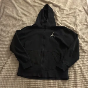 Svart Jordan hoodie med dragkedja - Svart hoodie från Jordan med klassisk logga på bröstet. Tröjan har hel dragkedja framtill, huva och två fickor. Tillverkad i mjuk bomullsblandning, perfekt för en sportig och avslappnad stil.