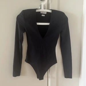 Svart långärmad bodysuit från Babaton - Säljer en svart bodysuit från Babaton med djup v-ringning och långärmad design. Snygg och stilren passform som sitter tight mot kroppen. Perfekt att styla med jeans eller kjol för en clean look. Materialet är mjukt och stretchigt för extra komfort. Shoulder pads 