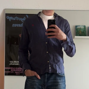 Blå skjorta från Polo Ralph Lauren - Snygg marinblå skjorta från Polo Ralph Lauren med klassisk button-down krage och röd broderad logga på bröstet. Skjortan är i bomull och har slim fit-passform med långa ärmar och knappar framtill. Perfekt för en clean och stilren look. S men passar M