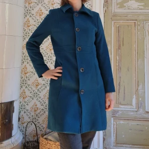 Blå kappa från LolaLiza stl 40(38) - Säljer en snygg petrolblå kappa från LolaLiza i storlek 40, något liten i storlek och passar bäst på storlek 38. Jag på bild har storlek 40 upptill, 42 över höfter. Kappan har klassisk krage, raka linjer och stängs med stora knappar framtill. Materialet känns mjukt och varmt, perfekt för kyligare dagar. Enkel och stilren design som passar till många outfits. Inga anmärkningar. Nyskick.