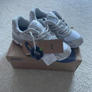 Vita Asics sneakers med mesh och mocka - Snygga vita Asics sneakers med detaljer i ljusgrått. Skorna har en sportig siluett, snörning och ovandel i mesh och mocka för extra andningsförmåga. Perfekta för dig som gillar stilrena och bekväma skor med chunky sula och klassisk Asics-logga på sidan.