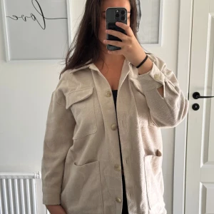 Beige overshirt från Nisan, strl 38 - Overshirt i beige från Nisan med stora bröstfickor, knappar framtill och klassisk krage. Jackan har lång ärm med knappdetalj vid ärmslut och är tillverkad i ett mjukt material som känns lite ulligt. Perfekt för lager-på-lager och chill stil.
