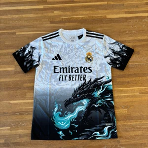 Real Madrid x Adidas draktröja M  | Vit med svart & turkos - .Snygg Real Madrid-fotbollströja från Adidas med ett unikt drakmotiv i svart och turkos på framsidan. Tröjan är vit med svarta detaljer, klassiska Adidas-ränder på axlarna och klubbmärke broderat på bröstet.  ✔️ Tillverkad i lätt och ventilerande material – perfekt till träning eller streetwear ✔️ Korta ärmar, regular fit ✔️ Aldrig använd  Storlek: M Skick: Ny / aldrig använd  Passar dig som vill ha en unik, limited edition-fotbollströja eller älskar Real Madrid. 
