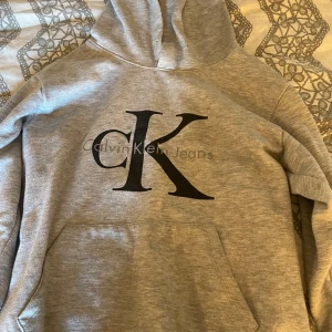 Grå hoodie från Calvin Klein Jeans - Snygg grå hoodie från Calvin Klein Jeans med stor svart CK-logga på bröstet. Tröjan har huva och en stor magficka framtill. Perfekt för en chill och stilren look. Mjuk och skön bomullsblandning som passar året runt.