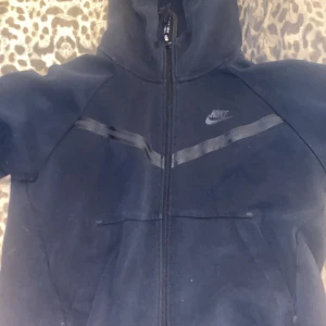 Svart Nike hoodie med dragkedja M - Svart hoodie från Nike i storlek M med hel dragkedja framtill, huva och två fickor. Snygga svarta detaljer över bröstet och klassisk Nike-logga på vänster sida. Perfekt för chill eller träning.