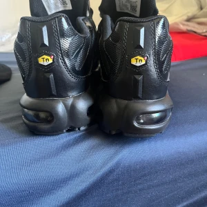 Nike Air Max Plus Tn Svarta Sneakers - Nike Air Max Plus Tn sneakers i helsvart färg med mesh och syntetpaneler, ikonisk Tn Air-dämpning och synliga luftkuddar i sulan. Snygg vågig design på ovandelen, snörning och rund tå. Perfekt för dig som gillar streetwear och sportig stil. INGEN ÅNGERRÄTT!