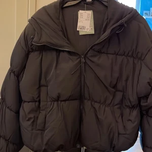 Svart pufferjacka med huva från H&M - Säljer en svart pufferjacka från H&M i storlek M. Jackan har en oversized look, stor huva och dragkedja framtill. Perfekt för kalla dagar med sin fluffiga och quiltade design. Enkel och stilren modell som håller dig varm hela vintern.