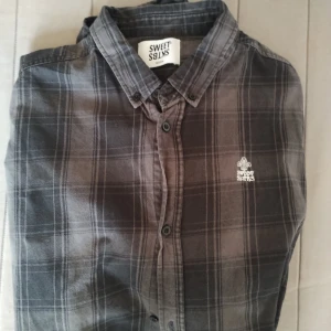 Grå rutig skjorta Sweet  M - Grå långärmad skjorta från Sweet SKBTS i storlek M. Skjortan har ett rutigt mönster i olika grå nyanser, klassisk krage och knappar framtill. Broderad logga på bröstet ger en extra detalj. Perfekt för dig som gillar avslappnad stil.