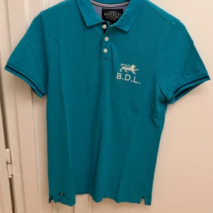 Turkos pikétröja från Bondelid - Snygg turkos pikétröja från Bondelid med klassisk krage, tre knappar och broderad logga med lejon och B.D.L. på bröstet. Tröjan har korta ärmar med svarta ränder vid ärmslut och är tillverkad i bomullspiké. Perfekt för en fräsch och sportig look. Storlek S.