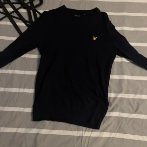 Mörkblå stickad tröja Lyle & Scott - Mörkblå stickad tröja från Lyle & Scott med klassisk gul broderad logga på bröstet. Tröjan har rund halsringning och långa ärmar, perfekt för lager-på-lager under kyliga dagar. Enkel och stilren design som funkar till det mesta.