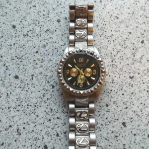 Dany Quartz silverfärgad klocka - Säljer en cool Dany Quartz klocka med svart urtavla och guldiga detaljer. Armbandet är i silverfärgad metall med unika mönstrade länkar och dekorativa symboler. Klockan har en rund form och markerad boett med pärldetaljer runt urtavlan.