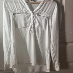 Vit långärmad henley med dragkedjor - Vit långärmad henley-skjorta med två bröstfickor med dragkedjor och knappslå framtill. Skjortan har en avslappnad passform och är tillverkad i ett lätt tyg som ger en luftig känsla. Perfekt för en clean och stilren look.