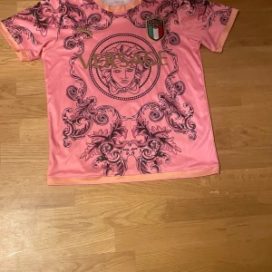 Rosa V3rsace x Puma Italien fotbollströja - Unik rosa fotbollströja med V3rsace-mönster, guldigt Puma-logga och Italiens landslagsemblem på bröstet. Tröjan har korta ärmar och är tillverkad i ett lätt, sportigt material. Stora V3rsace-loggan och Medusa-huvudet framtill ger en lyxig vibe.