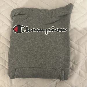 Grå hoodie från Champion - Grå hoodie från Champion med stor logga över bröstet och en mindre logga på ärmen. Klassisk känguruficka framtill och huva. Perfekt för chill dagar eller när du vill ha en sportig look.