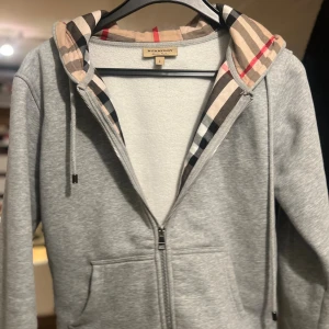 Grå Burberry hoodie med dragkedja - Säljer en grå hoodie från Burberry med klassiskt rutigt foder i huvan och broderad logga på bröstet. Tröjan har hel dragkedja, snörning i huvan och två fickor framtill. Perfekt för dig som vill ha en stilren och lyxig vardagslook.