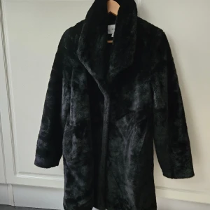 Svart pälsjacka från Vila, strl 36 - Mysig svart pälsjacka från Vila i storlek 36. Jackan har en bred krage, den är tillverkad i mjukt fuskpälsmaterial. Perfekt för kalla dagar när du vill ha en stilren och trendig look. Litet hål i fickan som kan sys. Använd 1 säsong