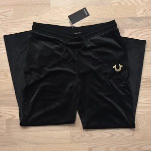 Svarta velourbyxor True Religion XL - Svarta mjukisbyxor i glansig velour från True Religion med guldfärgad logga på benet. Byxorna har rak passform, resår i midjan och snörning. Helt nya med tag. 