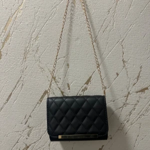Svart quiltad axelväska med kedja🤍 - Snygg svart axelväska med quiltat mönster och guldfärgad kedja som axelrem. Väskan har en stilren rektangulär form, gulddetalj framtill och ett fack med dragkedja inuti. Perfekt för att bära det viktigaste och ge din outfit en elegant touch.