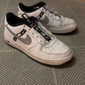 Nike Air Force 1 vita och grå sneakers - Säljer ett par Nike Air Force 1 sneakers i vitt skinn med grå swoosh och svarta detaljer. Skorna har svart innersula, svartvit Nike-logga på snörningen och svart sula. Klassisk låg modell med perforerad tå och tydliga Air-detaljer på sidan.