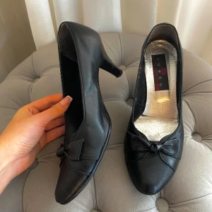 Svarta klackar  - Snygga svarta pumps från Romeo i storlek 38. Skorna har en klassisk klack, rund tå och är tillverkade i skinn. Framtill sitter en söt rosett som ger extra stilpoäng. Perfekta för dig som gillar tidlös design med en feminin touch.