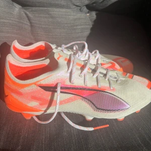 Puma vita  fotbollsskor Elite - Säljer ett par snygga Puma fotbollsskor i vitt och neonorange med svarta detaljer. Skorna har snörning och en låg, strömlinjeformad siluett med platt sula och dobbar för gräsplan. Ovandelen är tillverkad i syntetmaterial med coolt grafiskt mönster och Puma-logga på sidan.