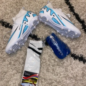 Vita och blå fotbollsskor strl 42 - Säljer ett par vita fotbollsskor med blå och svarta detaljer. Skorna har snörning, platt sula med dobbar och en högre strumpliknande krage för extra stöd. Materialet är syntet och designen är sportig med coola grafiska mönster. Perfekt för dig som vill sticka ut på planen.