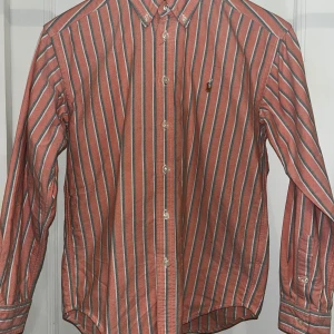 Randig skjorta från Ralph Lauren - Säljer en klassisk skjorta från Ralph Lauren i röd och vit randigt mönster med gröna detaljer. Skjortan har knappar hela vägen, button-down krage och lång ärm. Tillverkad i 100% bomull och har en broderad logga på bröstet.