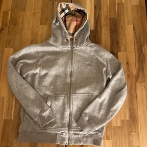 Grå Burberry hoodie med rutig huva - Säljer en grå hoodie från Burberry med dragkedja och klassiskt rutigt foder i huvan. Tröjan har två fickor fram, ribbade muddar och en snygg, stilren look. Perfekt för dig som gillar exklusiva streetwear-plagg med ikoniska detaljer.  Först till kvarn!.