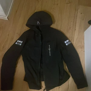 Svart Sail Racing pufferjacka med huva - Svart pufferjacka från Sail Racing med huva och dragkedja. Jackan har reflekterande loggor på båda ärmarna och bröstet, samt quiltad insida. Perfekt för kalla dagar och snygg stil.